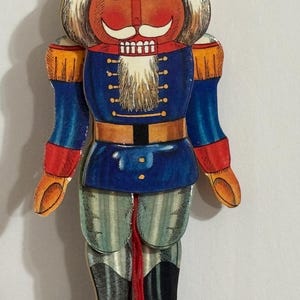 Op de afbeelding: Een vintage houten notenkraker speelgoed met een rood-oranje hoed, witte baard, blauw-wit gestreepte broek en rood-oranje laarzen. De notenkraker houdt een rood-oranje zwaard vast.