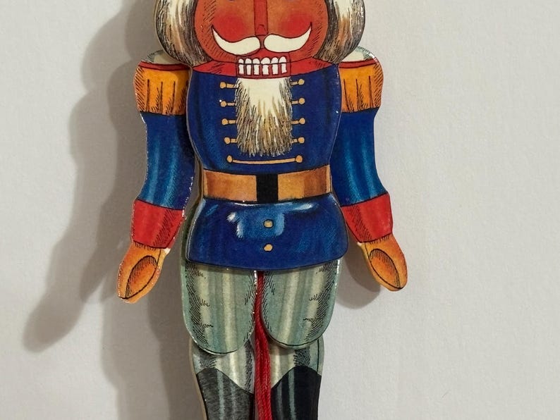 Op de afbeelding: Een vintage houten notenkraker speelgoed met een rood-oranje hoed, witte baard, blauw-wit gestreepte broek en rood-oranje laarzen. De notenkraker houdt een rood-oranje zwaard vast.