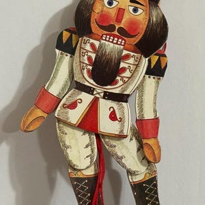 May include: A wooden nutcracker puppet with a red, white, and black painted design. The puppet is attached to a red string with a red ball at the end.
