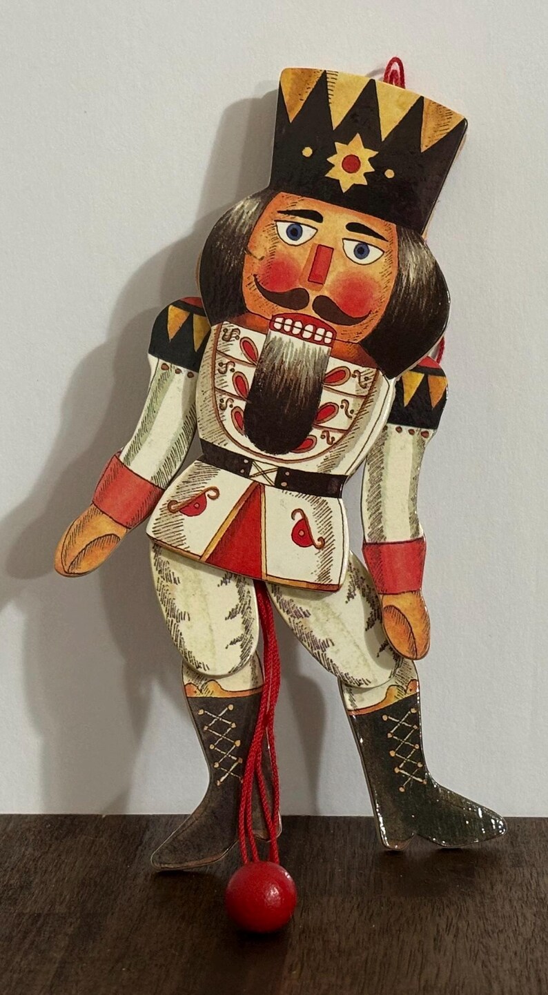 May include: A wooden nutcracker puppet with a red, white, and black painted design. The puppet is attached to a red string with a red ball at the end.