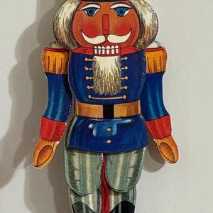 Op de afbeelding: Een houten notenkraker ornament met een rood, wit en blauw uniform. De notenkraker heeft een witte baard, een zwarte hoed met een wit hakbijl ontwerp en een rode bal aan het einde van een touw.
