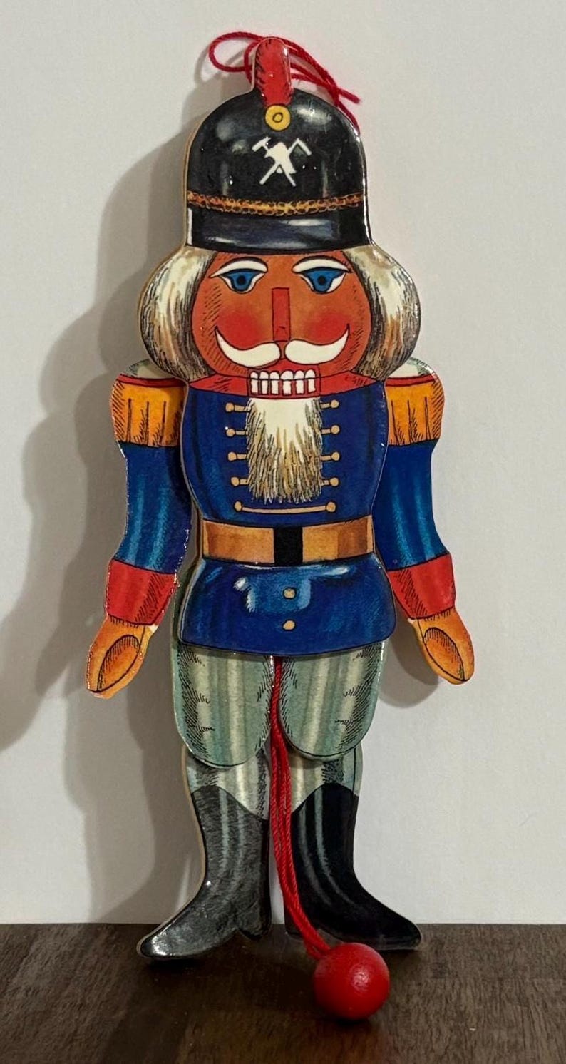 Op de afbeelding: Een houten notenkraker ornament met een rood, wit en blauw uniform. De notenkraker heeft een witte baard, een zwarte hoed met een wit hakbijl ontwerp en een rode bal aan het einde van een touw.
