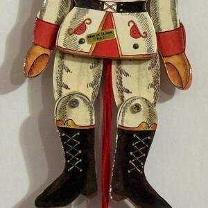 May include: A vintage wooden pull-string toy soldier with red, white, and black details. The soldier is wearing a white uniform with red trim, black boots, and a red sash. The toy has a pull string attached to its legs, and the text "MADE IN TAIWAN R.G.C." is printed on the front.