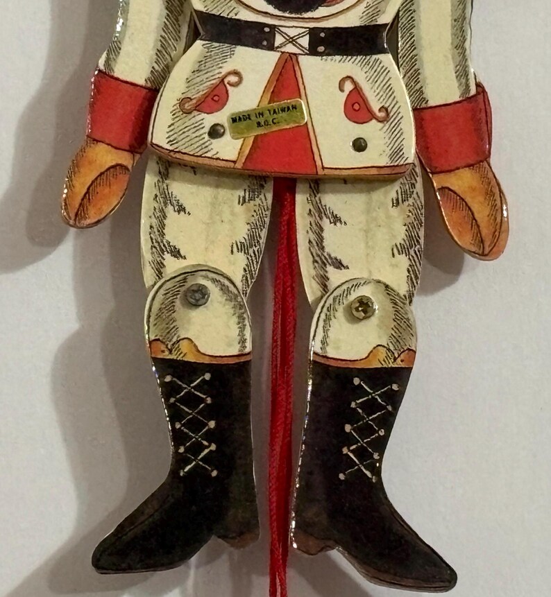 May include: A vintage wooden pull-string toy soldier with red, white, and black details. The soldier is wearing a white uniform with red trim, black boots, and a red sash. The toy has a pull string attached to its legs, and the text "MADE IN TAIWAN R.G.C." is printed on the front.