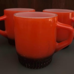 Puede incluir: Tres tazas de vidrio naranja con interiores blancos y fondos negros. Las tazas tienen una forma redondeada y un asa a cada lado.