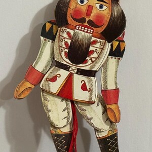 May include: A wooden nutcracker doll with a red, white, and black painted design. The doll is wearing a black hat with a gold star and has a red and white painted face with a black mustache and beard. The doll is holding a red string.