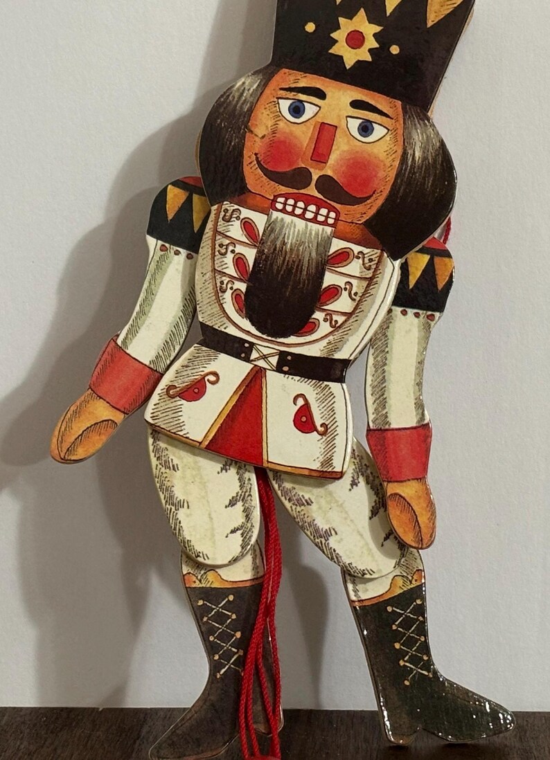 May include: A wooden nutcracker doll with a red, white, and black painted design. The doll is wearing a black hat with a gold star and has a red and white painted face with a black mustache and beard. The doll is holding a red string.