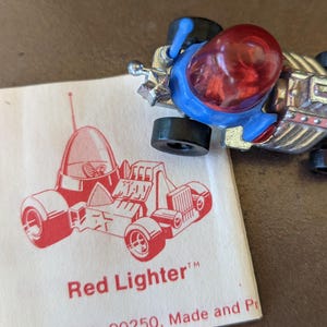 May include: A vintage toy car with a red bubble-shaped cockpit and blue accents. The car is silver with black wheels. A white card with a red illustration of the car and the text "Red Lighter" is next to the car.