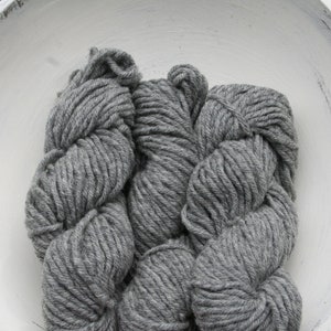May include: Three skeins of gray yarn are arranged inside a white, distressed wooden bowl. The yarn appears thick and textured, with a slightly heathered appearance. The bowl has a rustic, farmhouse aesthetic.