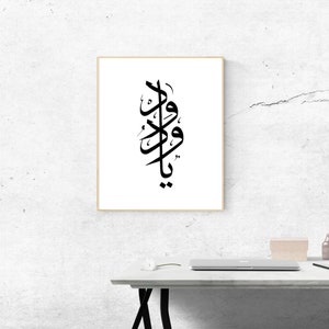 May include: Black and white calligraphy art print with the Arabic word "Ya Wadood" in a decorative style. The print is framed in a light wood frame and is hanging on a white wall.