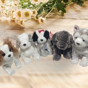 May include: Five plush dog toys in various breeds and colors are arranged on a wooden surface with white flowers. The dogs include a Cavalier King Charles Spaniel, a white and gray terrier, a black and white dog with a red bow, a gray dog, and a gray and white husky.