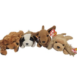 May include: Four plush dog toys in various colors and breeds are displayed. The dogs include a brown poodle, a black and white spotted dog, a brown dachshund, and a tan golden retriever. Each dog has a soft, plush texture.