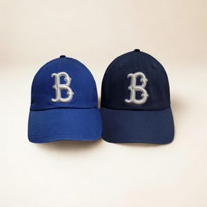 May include: Two baseball caps, one in royal blue and the other in navy blue. Both caps feature a white embroidered "B" on the front. The caps have a curved brim and a fabric strap closure on the back.