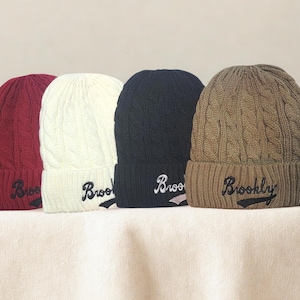 May include: Four cable-knit beanies in burgundy, cream, navy, and tan. Each beanie features a folded brim and the embroidered word "Brooklyn" in a script font. The hats are displayed on a neutral surface.