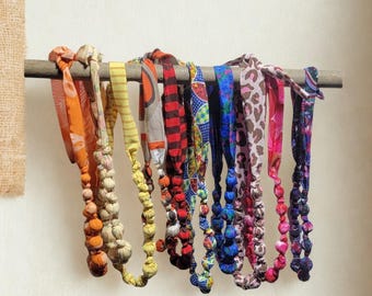 Op de afbeelding: Een verzameling kleurrijke stoffen kettingen die aan een houten staaf hangen. De kettingen hebben verschillende patronen en kleuren, waaronder oranje, geel, rood, blauw en roze. Elke ketting heeft een reeks met stof bedekte kralen.