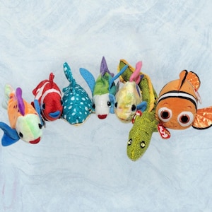 May include: A collection of seven plush fish toys featuring various colors and patterns, including a bright orange clownfish, a red and white striped fish, a blue spotted fish, a green and white fish, and a long, textured green eel-like plush.