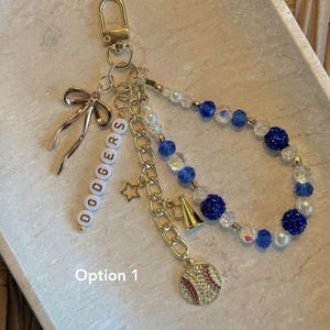 May include: A gold-toned keychain featuring a lobster clasp, a metal bow charm, a strand of white beads spelling DODGERS, a gold chain with a star, a megaphone charm, a baseball charm, and a loop of blue, white, and clear faceted beads. Text: Option 1.