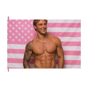 May include: A flag featuring a pink and white striped pattern with a pink canton containing white stars. The flag serves as a backdrop for a person with a muscular build, a tattoo on the right arm, and a thin silver chain necklace around the neck.