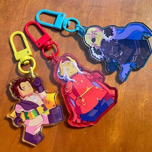 May include: Three acrylic keyrings featuring cartoon characters. One keyring has a yellow clasp and a character in a purple and gold outfit. Another has a red clasp and a character in a red outfit. The third has a blue clasp and a character in a dark outfit.