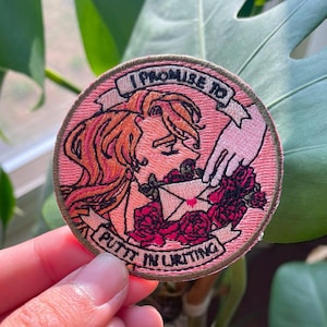 May include: Pink embroidered patch featuring a woman with long blonde hair, a hand holding a letter, and flowers. The text on the patch reads "I promise to put in writing".