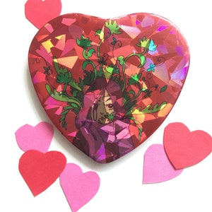 以下が含まれることがあります： ホログラフィック仕上げの赤いハート型の缶には、頭から植物が生えている人のカラフルなイラストが描かれています。缶は赤とピンクの紙のハートで囲まれています。