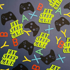 May include: A collection of colorful confetti featuring the phrase "Eat Sleep Game" in green and black, along with various letters and game controller shapes in black, red, yellow, blue, and green.