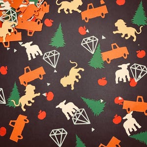 May include: A collection of colorful paper cutouts scattered on a dark background. The cutouts include orange trucks, yellow lions, white sheep, green trees, red apples, and white diamond shapes. The overall theme suggests a playful, whimsical design.