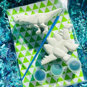 May include: A dinosaur-themed paint kit featuring two white plaster dinosaur figures, a blue paintbrush, and three small paint pots with blue and teal paint. The kit is packaged in a clear plastic bag with a green and white triangle pattern.