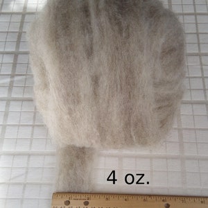 May include: A close-up of a 4 ounce bundle of light gray wool roving. The wool is arranged in a loose, fluffy pile. A ruler is visible in the bottom of the image.
