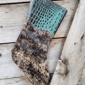 May include: A Christmas stocking with a cowhide body and a turquoise faux-crocodile leather cuff. The stocking is brown, black, and tan with a leather loop for hanging. The cuff is a dark teal color with a brown border.