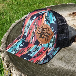 May include: A black and red trucker hat with a colorful southwestern print featuring cacti and horses. The hat has a black mesh back and a brown leather patch with the text "Just Rope It".