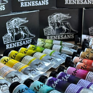 May include: A selection of Renesans watercolour paint tubes in diverse shades, such as yellow, green, blue, and purple. The tubes are neatly arranged, with black boxes displaying a lion logo visible in the background.