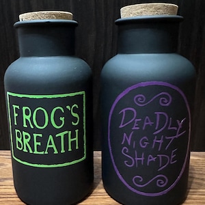 May include: Two black apothecary jars with cork stoppers. One jar has a green label with "FROG'S BREATH" in white. The other jar has a purple label with "DEADLY NIGHT SHADE" in white. The jars are on a dark wooden surface.