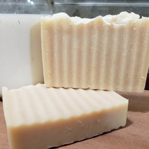 May include: Two bars of handmade soap, one on top of the other. The soap is a light beige color and has a rippled texture. The soap is sitting on a wooden surface.
