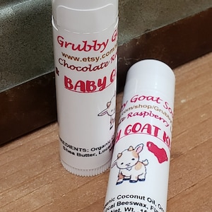 May include: Two white lip balm tubes with red and black labels. The left tube has a cartoon goat and the text "Grubby Goat Chocolate Raspberry Baby Goat Kiss". The right tube has a cartoon goat and the text "Baby Goat Soap Raspberry Goat Kiss". The text on the right tube reads "Organic Coconut Oil, Organic Local Beeswax, Flavor Oil. Net. Wt. .15 oz."