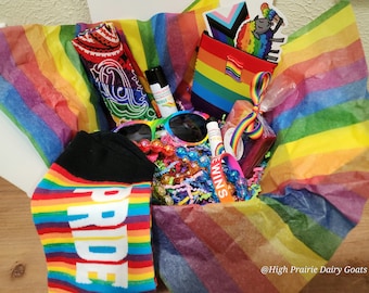 May include: A rainbow-themed gift box filled with various items, including socks, a bandana, sunglasses, lip balm, a soap bar, and a rainbow-colored pouch. The box is lined with rainbow-striped tissue paper and includes a small sticker with a cartoon sheep. The text "@High Prairie Dairy Goats" is visible at the bottom of the image.