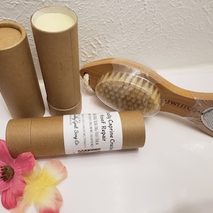 May include: Two brown cardboard tubes with white labels that read "HooF Repair Naturally Healing Foot Balm -  Made with 100% Caprine Grade -  Body Goat Soap Co.". One tube is open and contains a white balm. A wooden foot brush with a bristle brush on one end and a metal foot file on the other end. The brush is wrapped in plastic.