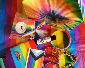 May include: A collection of Pride-themed items on a rainbow striped background. Includes a rainbow-colored gift box, a rainbow bracelet, a can cooler, a sticker, a lip balm, a rainbow-colored ribbon, and a package of rainbow-colored soap.