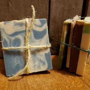 May include: Two bars of handmade soap, one wrapped in twine and the other wrapped in green string. The soap on the left is blue and white marbled. The soap on the right is a stack of four bars with different colors and patterns.