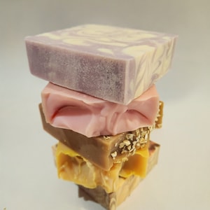 May include: A stack of four handmade soap bars. The top bar is purple and white with swirls. The second bar is pink with swirls. The third bar is brown with oatmeal flakes. The bottom bar is yellow with swirls.