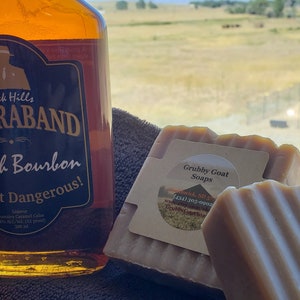 May include: Two bars of Grubby Goat Soaps, one brown and one beige, with a label that reads "Grubby Goat Soaps" and includes a phone number and website address. The soaps are sitting on a brown towel next to a bottle of Black Hills Raband Bourbon.