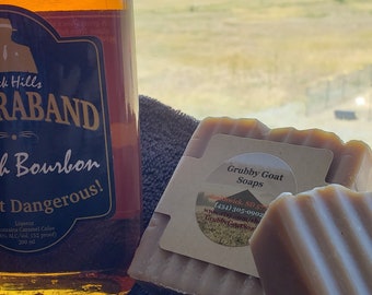 May include: Two bars of Grubby Goat Soaps, one brown and one beige, with a label that reads "Grubby Goat Soaps" and includes a phone number and website address. The soaps are sitting on a brown towel next to a bottle of Black Hills Raband Bourbon.