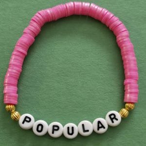 May include: Pink beaded bracelet with gold accents and white letter beads spelling "POPULAR".