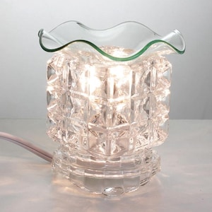 May include: A clear, illuminated glass essential oil diffuser with a square, textured body and a wavy-edged dish. The base is round and the diffuser is plugged into a white electrical cord. The light shines through the glass.
