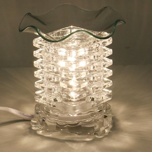 May include: A clear glass electric wax warmer featuring a square, ribbed body with a ruffled top dish. The lamp is illuminated from within, casting a bright glow through the textured glass structure, and includes a white power cord extending from the base.
