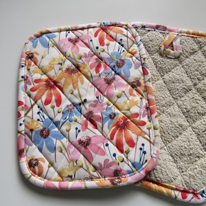 May include: Floral print pot holder with a quilted design. The pot holder features a white background with colorful flowers in shades of red, orange, pink, blue, and yellow. The reverse side is a beige terry cloth material.