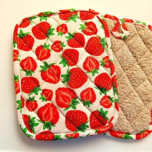 Puede incluir: Un par de agarraderas de olla acolchadas. Una es blanca con un patrón de fresas rojas y la otra es beige. Ambas tienen un lazo para colgar.