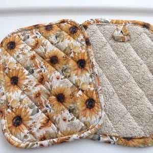May include: Two square pot holders featuring a sunflower pattern with yellow petals and dark brown centers on a cream background. One side displays the floral print with diagonal quilting, while the other side shows a textured beige fabric.