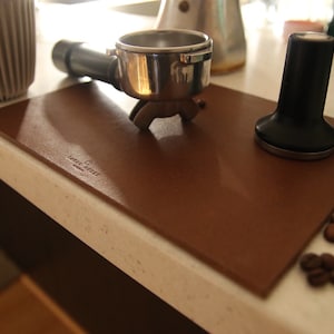 Könnte beinhalten: Eine braune Leder-Kaffeematte mit dem aufgedruckten Text "COFFEE SOCIETY". Auf der Matte liegen ein silberner Siebträger, ein schwarzer Tamper und Kaffeebohnen.