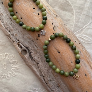 Könnte beinhalten: Zwei grüne Jade-Armbänder mit goldenen Blatt-Anhängern. Die Armbänder liegen auf einer Holzoberfläche.